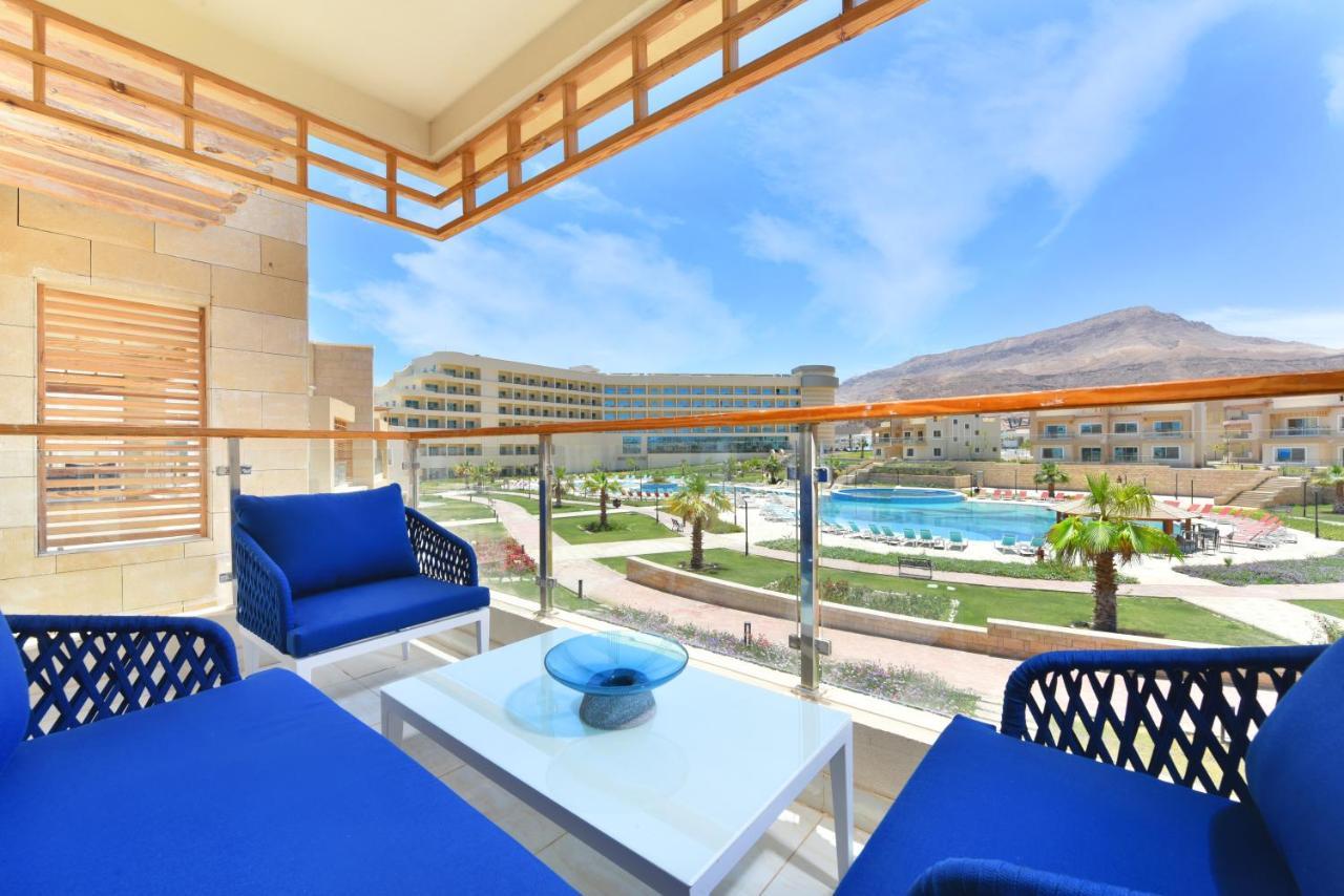 Tolip Resort El Galala Majestic Ain Sukhna Exterior photo The photo showcases a balcony view featuring two blue chairs and a table with a decorative bowl. The balcony overlooks a landscape that includes a swimming pool and manicured lawns. In the background, there are multiple buildings and palm trees, all 