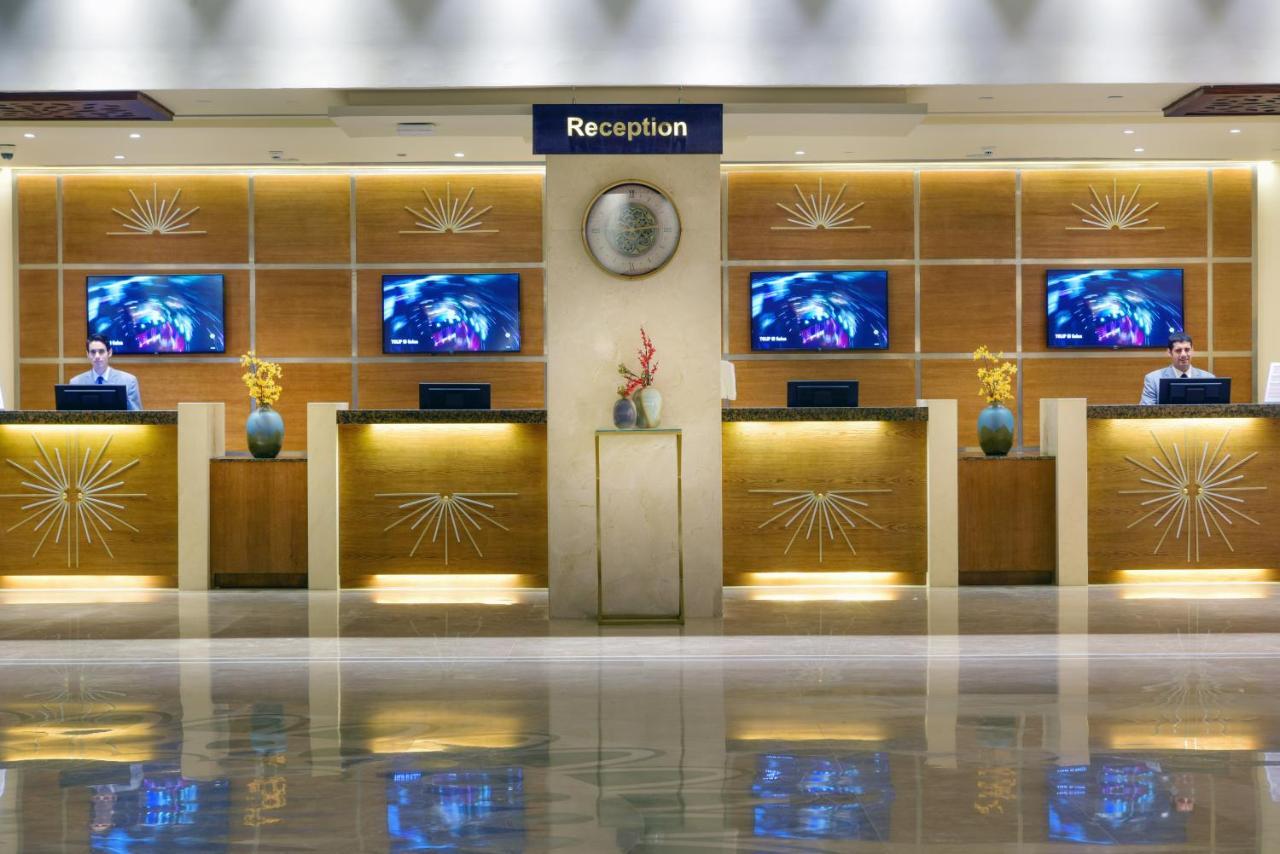 Tolip Resort El Galala Majestic Ain Sukhna Exterior photo The image shows a hotel reception area. There are several reception desks lined up against a wall with decorative paneling. Each desk has a staff member, and above the desks, there are screens displaying information. A clock is visible on the wall, a