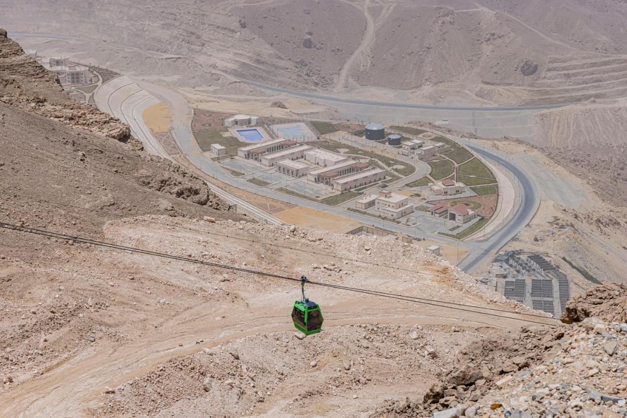 Tolip Resort El Galala Majestic Ain Sukhna Exterior photo The photo shows a mountainous landscape with a steep, dry terrain. In the foreground, there is a green cable car or gondola traveling along a cable. Below, a complex of buildings and roads can be seen, likely part of a resort or recreational area. Th