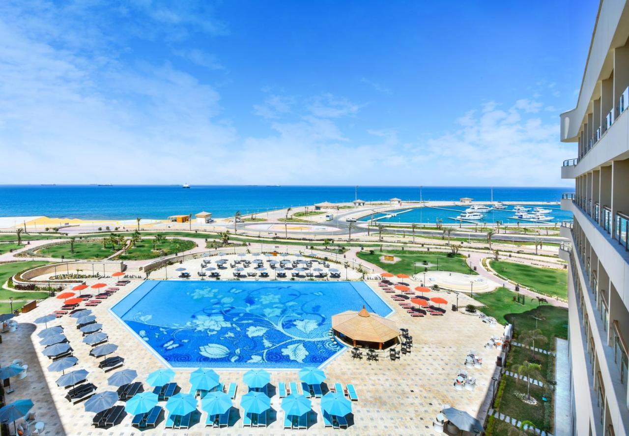 Tolip Resort El Galala Majestic Ain Sukhna Exterior photo The photo shows a beautiful outdoor view of a resort area featuring a large swimming pool with a decorative underwater design. Surrounding the pool are numerous sun loungers and umbrellas in a variety of colors. In the background, there is a scenic c