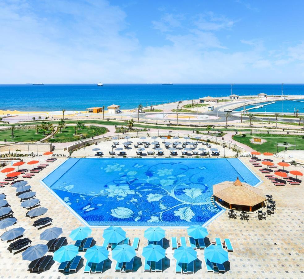 Tolip Resort El Galala Majestic Ain Sukhna Exterior photo The photo features a large swimming pool with a decorative design on its surface, surrounded by sun loungers and umbrellas. In the background, there is a view of the sea, with a clear blue sky and a few scattered clouds. The area is well-maintained, 