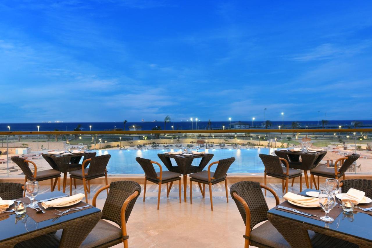 Tolip Resort El Galala Majestic Ain Sukhna Exterior photo The photo shows an outdoor dining area with several neatly arranged tables and chairs. The tables are elegantly set with cutlery and plates, suggesting a fine dining experience. In the background, there is a large swimming pool that reflects the blue